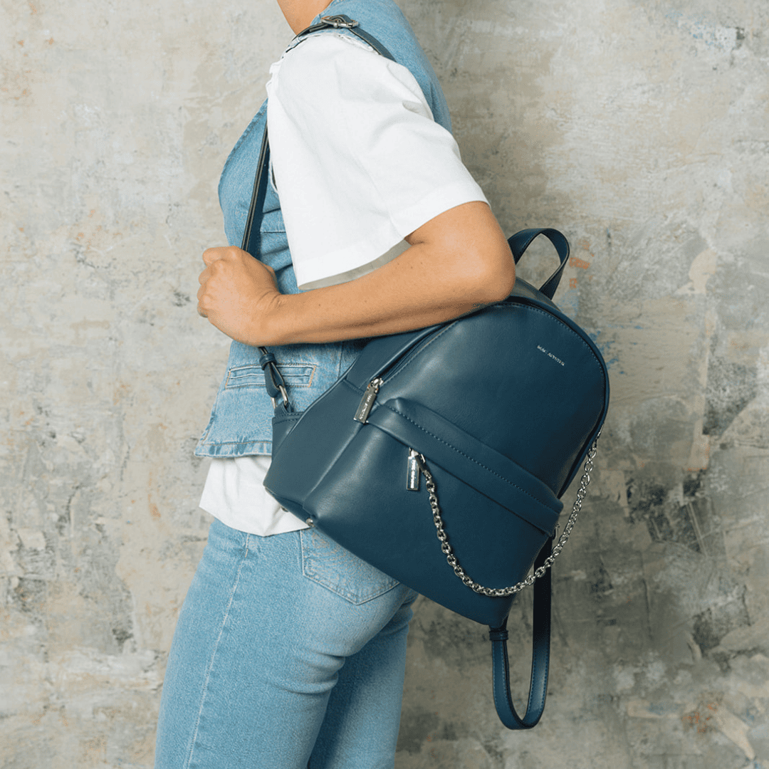 Mannequin de profil portant un sac à dos bleu marine en cuir avec détails métalliques et chaîne décorative. Tenue en jean et t-shirt blanc, fond texturé effet béton.