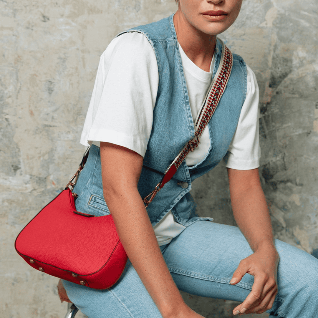une femme vêtue d'un ensemble en jean et d'un t-shirt blanc porte un sac rouge vif à la bandoulière tressée. L'association du denim et de l'accessoire audacieux crée un style à la fois moderne et décontracté