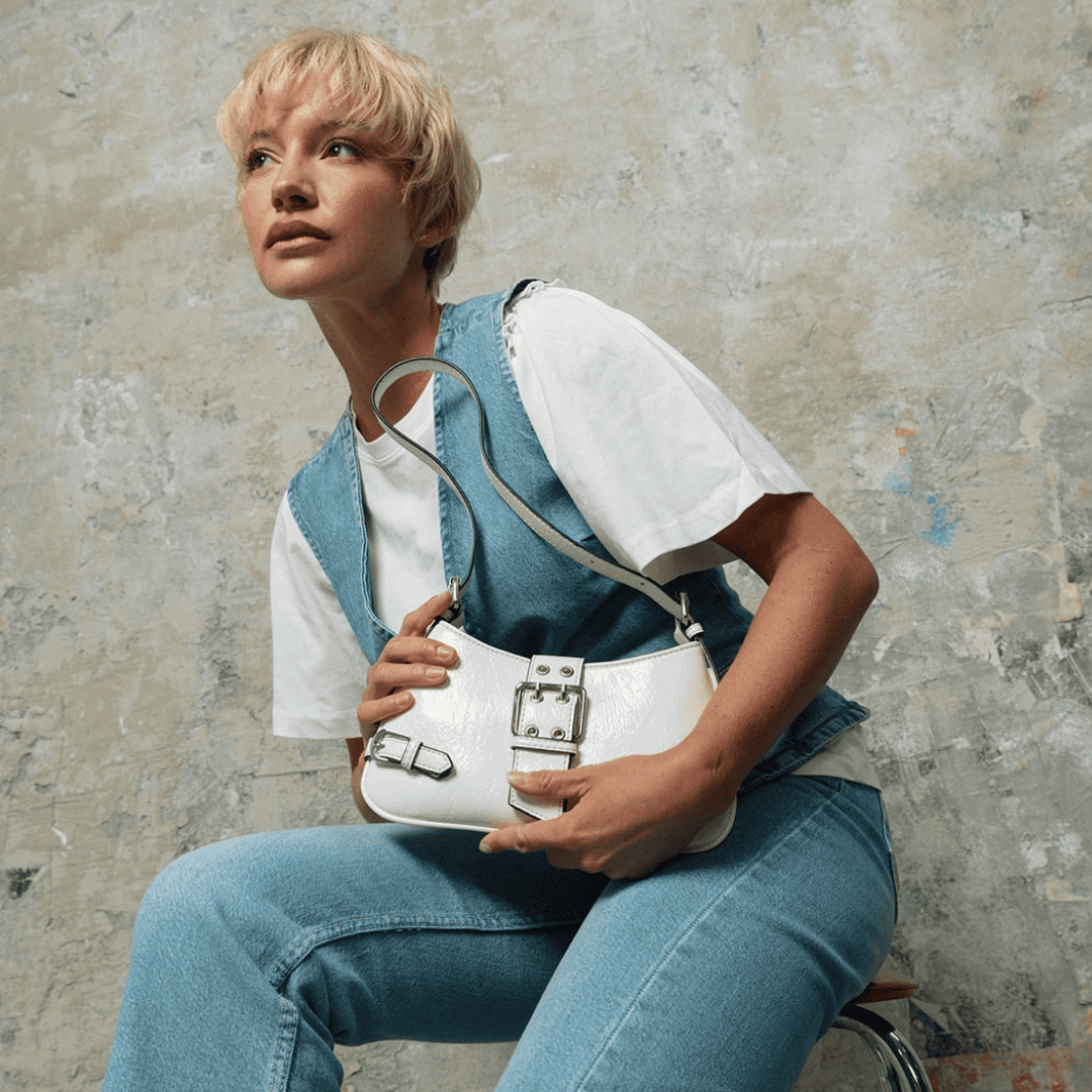 Mannequin en total look denim et t-shirt blanc, tenant un sac à main blanc effet vinyle avec détails de boucles argentées et une anse réglable. Ambiance moderne et audacieuse sur fond texturé effet béton.