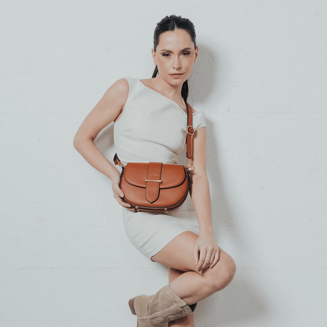 Une femme au look sophistiqué pose avec assurance devant un fond blanc texturé. Elle porte une robe blanche minimaliste, mettant en valeur son allure élégante. À son épaule, un sac en cuir camel au design structuré, orné d'une boucle raffinée et d'une bandoulière ajustable. Ses bottes en daim beige complètent son style chic et intemporel, créant un contraste harmonieux avec les tons naturels de son accessoire.