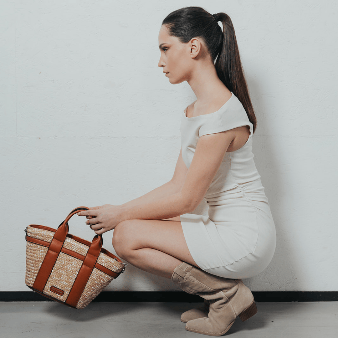 Une femme élégante, vêtue d'une robe blanche minimaliste, pose avec grâce en tenant un sac en raphia tressé aux détails en cuir camel. Son allure sophistiquée est accentuée par sa posture et son regard tourné vers le haut. Un mélange parfait entre chic et naturel, idéal pour une touche estivale raffinée.