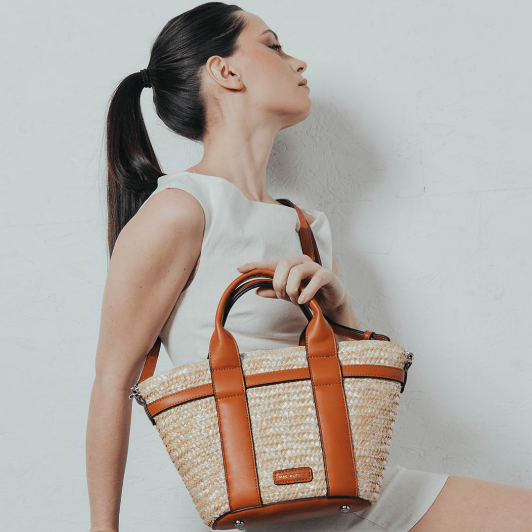 Une femme élégante, vêtue d'une robe blanche minimaliste, pose avec grâce en tenant un sac en raphia tressé aux détails en cuir camel. Son allure sophistiquée est accentuée par sa posture et son regard tourné vers le haut. Un mélange parfait entre chic et naturel, idéal pour une touche estivale raffinée.