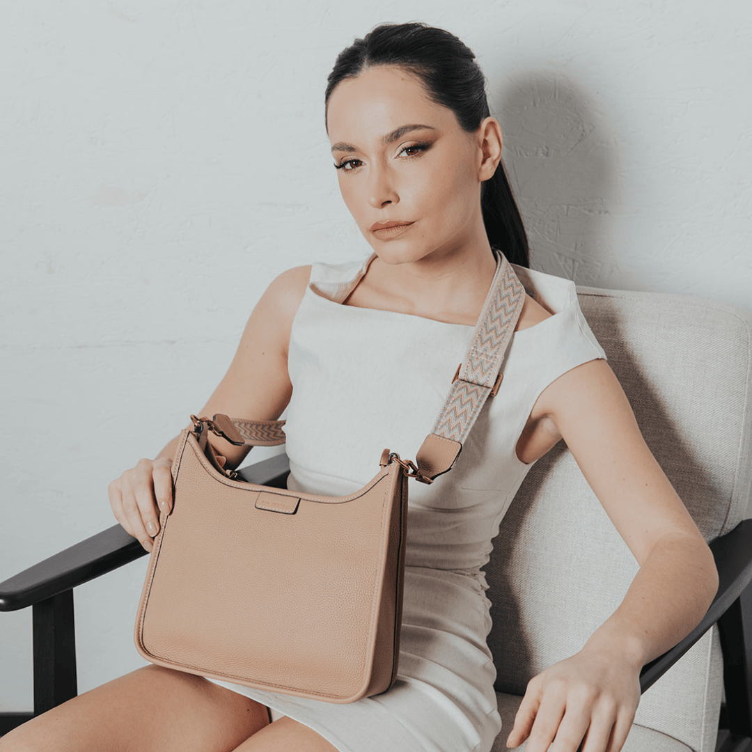 une femme au regard intense, coiffée d'une file de cheval soignée, porte une robe blanche épurée et un sac beige avec une large bandoulière à motif géométrique. Assise sur un fauteuil en tissu, elle incarne un style chic et intemporel