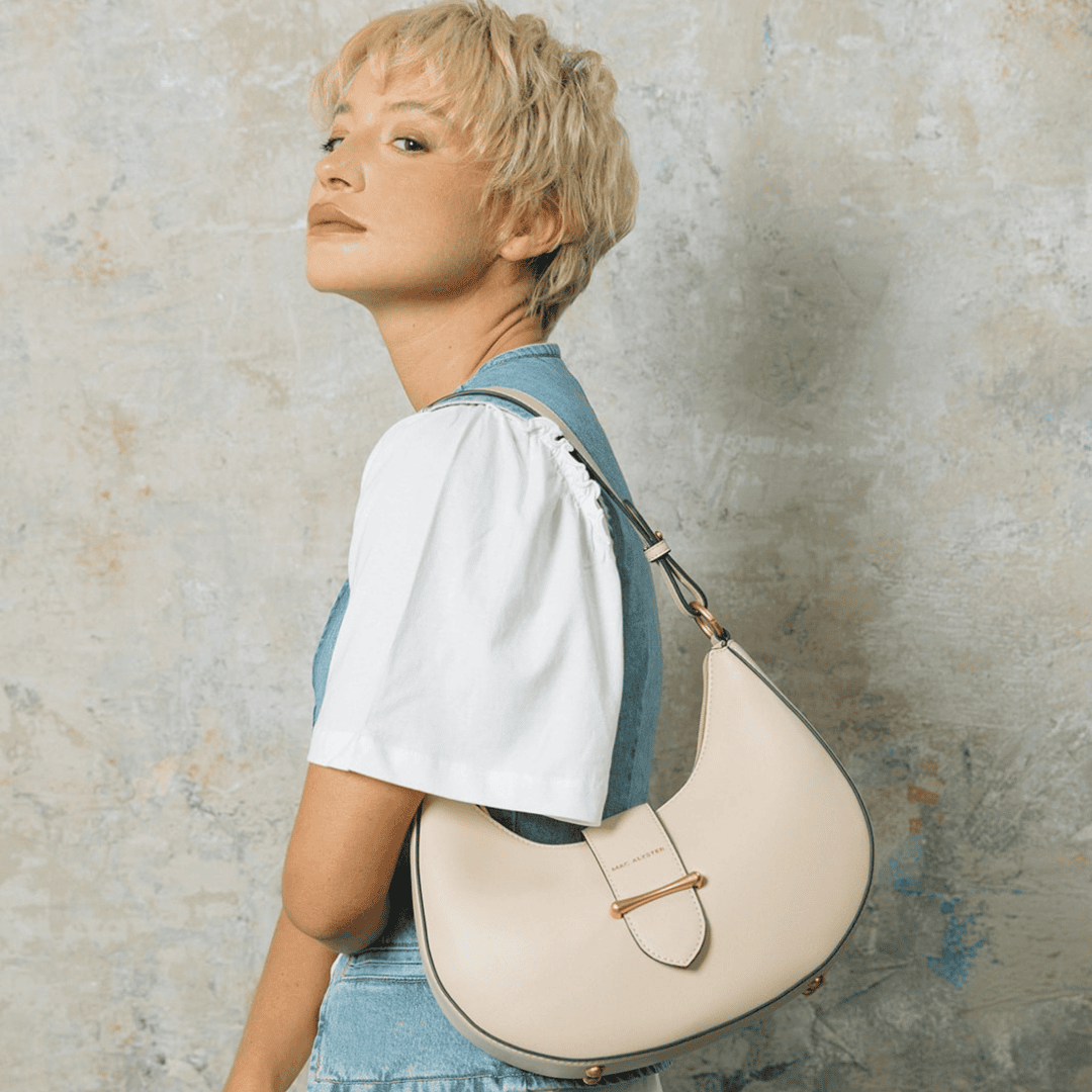 Mannequin en tenue denim et t-shirt blanc portant un sac à main crème en forme de demi-lune, avec une anse réglable et un détail métallique doré sur le rabat. Look minimaliste et élégant sur fond texturé effet béton.