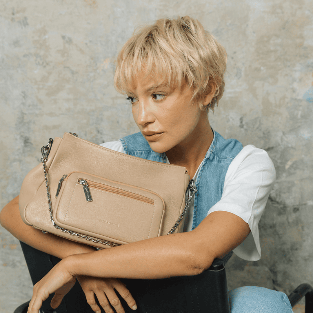 Mannequin assis tenant un sac à main en cuir beige avec poche zippée à l’avant et chaîne en métal. Look décontracté en jean et t-shirt blanc, fond texturé effet béton.