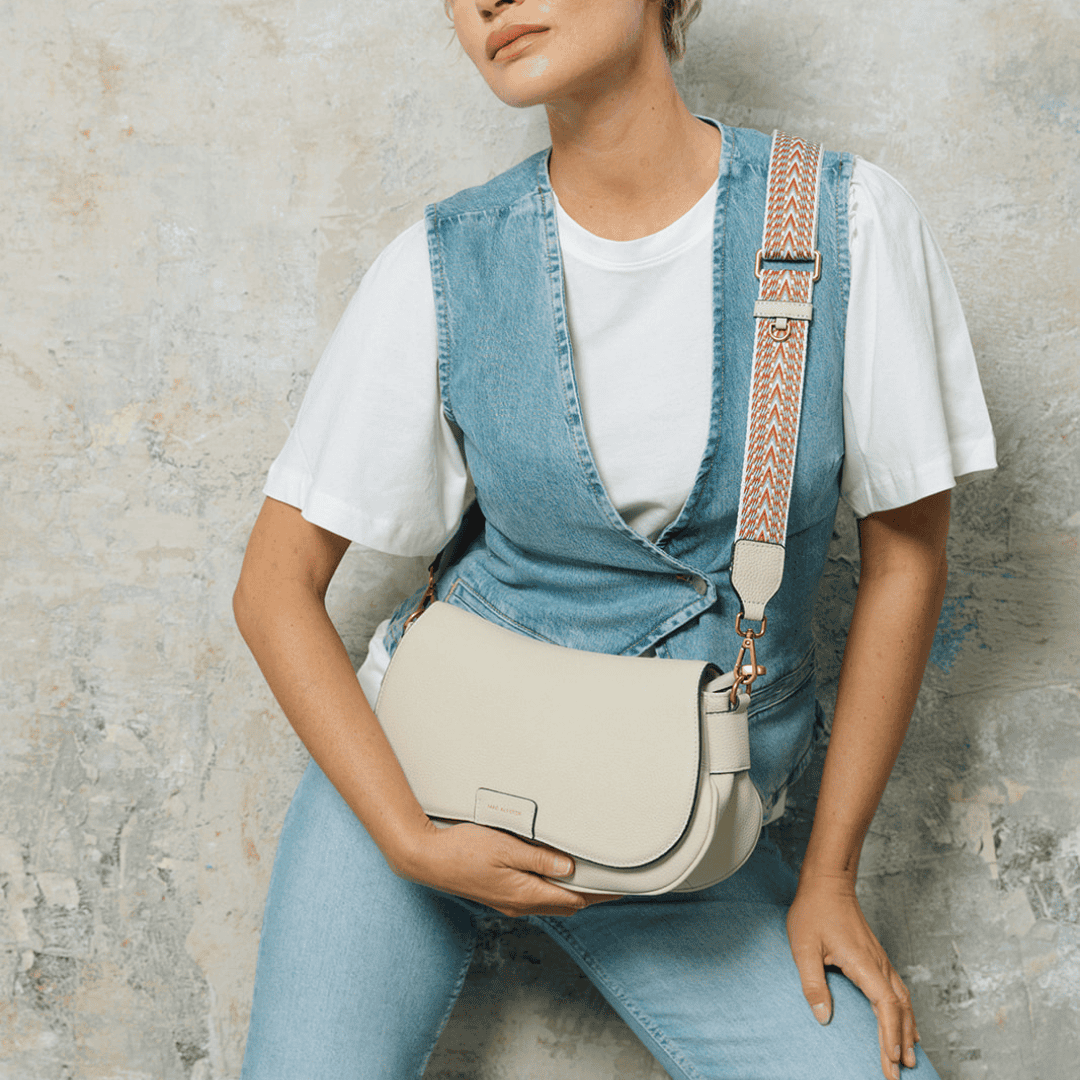 Mannequin en tenue denim et t-shirt blanc portant un sac à main crème à rabat, avec une bandoulière large et tissée aux motifs colorés. Style casual chic sur fond texturé effet béton.